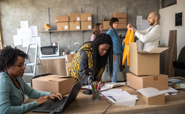 A woman working on a laptop while three others pack items for shipment in a room filled with cardboard boxes, illustrating eCommerce operations.
